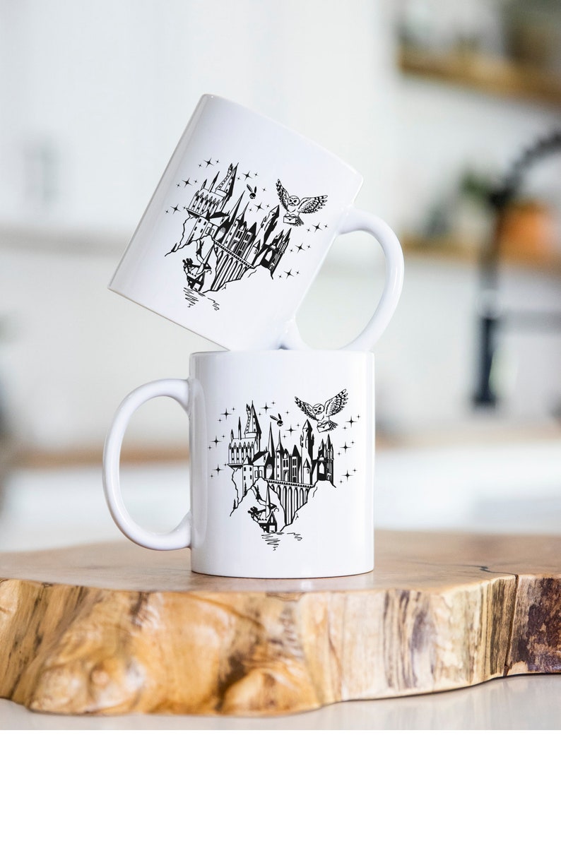 Puede incluir: Dos tazas de cer&aacute;mica blanca con una ilustraci&oacute;n negra de un castillo, un b&uacute;ho y estrellas. Las tazas est&aacute;n apiladas sobre una superficie de madera. El dise&ntilde;o tiene un tema de fantas&iacute;a.