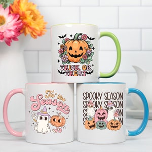 May include: Three white ceramic mugs with colorful interiors and handles. Each mug features a Halloween-themed design: a jack-o'-lantern with text "Trick or Treat", a ghost and pumpkin with text "Tis the Season", and a group of pumpkins and ghosts with text "Spooky Season".