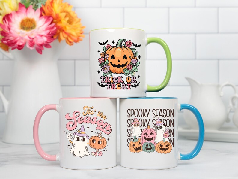 May include: Three white ceramic mugs with colorful interiors and handles. Each mug features a Halloween-themed design: a jack-o'-lantern with text "Trick or Treat", a ghost and pumpkin with text "Tis the Season", and a group of pumpkins and ghosts with text "Spooky Season".