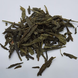 May include: A pile of loose leaf green tea, with long, thin, dark green leaves.