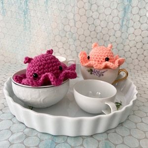 May include: Two crocheted octopus plushies, one pink and one peach, sitting in teacups on a white tray.