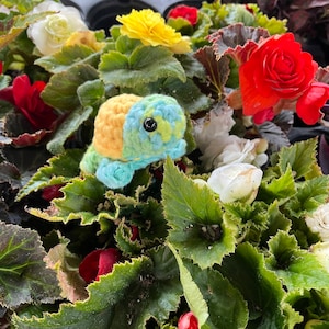 Puede incluir: Un juguete de tortuga de crochet con un esquema de colores amarillo, verde y azul. La tortuga está sentada en una cama de hojas verdes y flores.