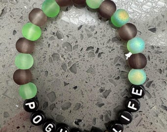 Puede incluir: Una pulsera de cuentas con una mezcla de cuentas verdes, marrones e iridiscentes. La pulsera incluye cuentas de letras blancas y negras que deletrean "POGUE 4 LIFE" y cuentas doradas. Las cuentas están ensartadas para formar un círculo.