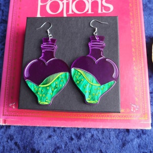 May include: A pair of purple heart-shaped bottle earrings with green and gold liquid inside. The earrings are made of acrylic and have silver hooks.