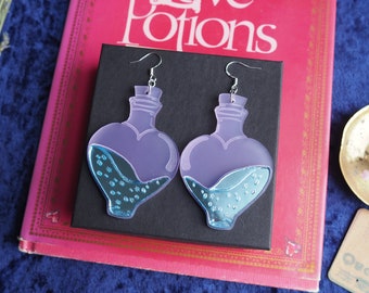 Peut inclure: Une paire de boucles d'oreilles en forme de bouteille de cœur avec une teinte violette clair et un liquide bleu à l'intérieur. Le liquide contient de petites bulles blanches. Les boucles d'oreilles sont suspendues à des crochets argentés.