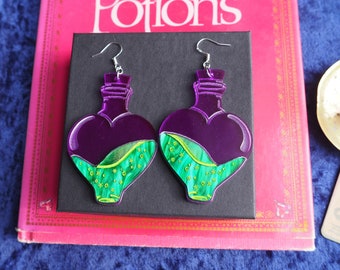 Peut inclure: Une paire de boucles d'oreilles en forme de cœur en bouteille violette avec un liquide vert et or à l'intérieur. Les boucles d'oreilles sont en acrylique et ont des crochets argentés.