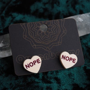 Puede incluir: Un par de pendientes de corazón blanco con la palabra "NOPE" impresa en rojo en cada pendiente. Los pendientes se muestran en una tarjeta negra con un patrón decorativo.