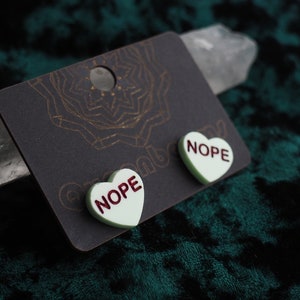Puede incluir: Un par de pendientes en forma de corazón con la palabra "NOPE" impresa en rojo sobre un fondo blanco. Los pendientes están en una tarjeta negra con un diseño de mandala dorado.