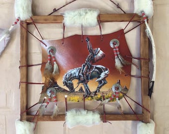 May include: A framed Native American-themed artwork. The artwork features a painting of a Native American on horseback, set against a red and orange background. The frame is made of wood and adorned with feathers, fur, and decorative elements.