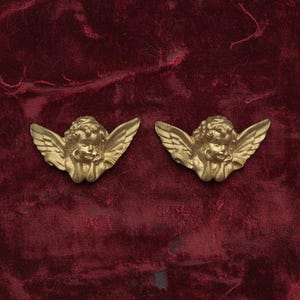 May include: Two gold cherub figurines with detailed wings and facial features. They are set against a deep red velvet background. The cherubs are a classic decorative element. The contrast between the gold and red creates a striking visual effect.