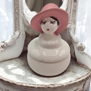 May include: A white ceramic trinket box shaped like a woman wearing a pink hat. The woman's face has closed eyes, black hair, and red lips. The box is on a white, ornate table.