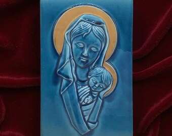 May include: A rectangular, blue ceramic plaque featuring a relief of the Virgin Mary holding the baby Jesus. The figures are in a lighter shade of blue, with a gold halo. The background is a deep red.