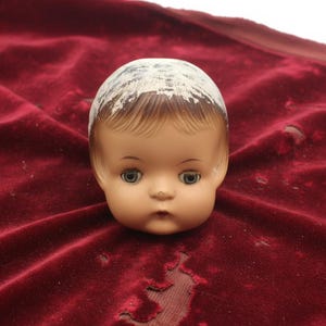 May include: A doll head with a light peach complexion, painted blue eyes, and brown hair. The top of the head features a white painted area with visible brush strokes. The doll has a small nose, defined eyebrows, and a neutral, closed-mouth expression.