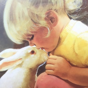 May include: A young girl with blonde hair is gently kissing a white rabbit on the nose. The girl is wearing a yellow shirt and pink pants. The rabbit is looking up at the girl with its eyes closed.