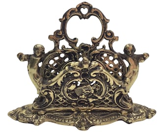 May include: A gold-tone metal letter holder with ornate details and two cherub figures on the sides. The holder has a decorative, openwork design.