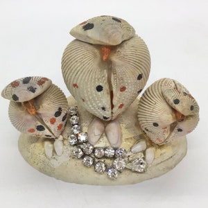 May include: Three small owl figurines made from seashells with black and orange accents. The owls are arranged on a base with a rhinestone necklace.