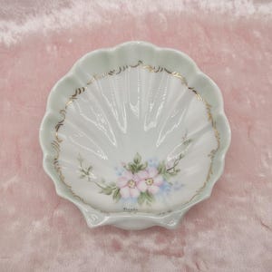 May include: A seashell-shaped trinket dish with a scalloped edge. The dish is white with a pale green rim and gold trim. It features a hand-painted floral design with pink and blue flowers.