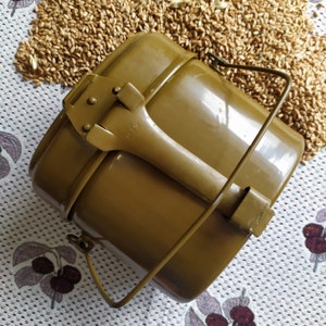 May include: A green metal camping mess kit with a folding handle and a lid. The kit is sitting on a surface covered in wheat kernels.