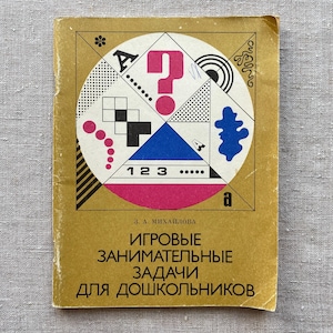 Può includere: Un libro per bambini vintage con una copertina dorata con un design geometrico con un punto interrogativo rosa, forme in bianco e nero e i numeri 1, 2 e 3. Il titolo è in russo.