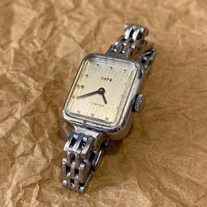 May include: A silver wristwatch with a square face and a silver metal band. The watch face is white with black hands and black markings. The watch face has the text "ЗАРЯ 17 КАМНЕЙ" printed on it.