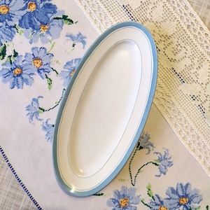 May include: A white oval serving platter with a light blue trim. The platter is on a white tablecloth with a blue floral pattern.