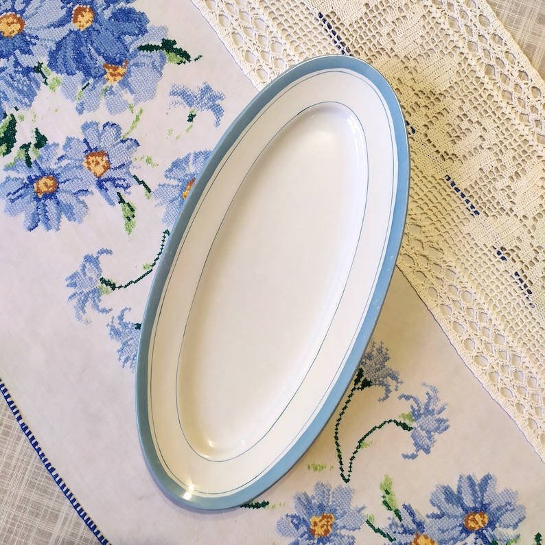 May include: A white oval serving platter with a light blue trim. The platter is on a white tablecloth with a blue floral pattern.