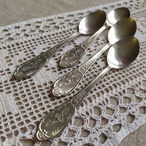 May include: Four silver spoons with ornate floral designs on a white crocheted doily.