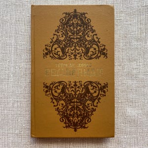 Op de afbeelding: Een vintage boek met een bruine kaft en sierlijke bruine bloemmotieven. De titel en de naam van de auteur staan in gouden letters. Het boek is gesloten en lijkt in goede staat te verkeren, wat suggereert dat het goed bewaard is gebleven.