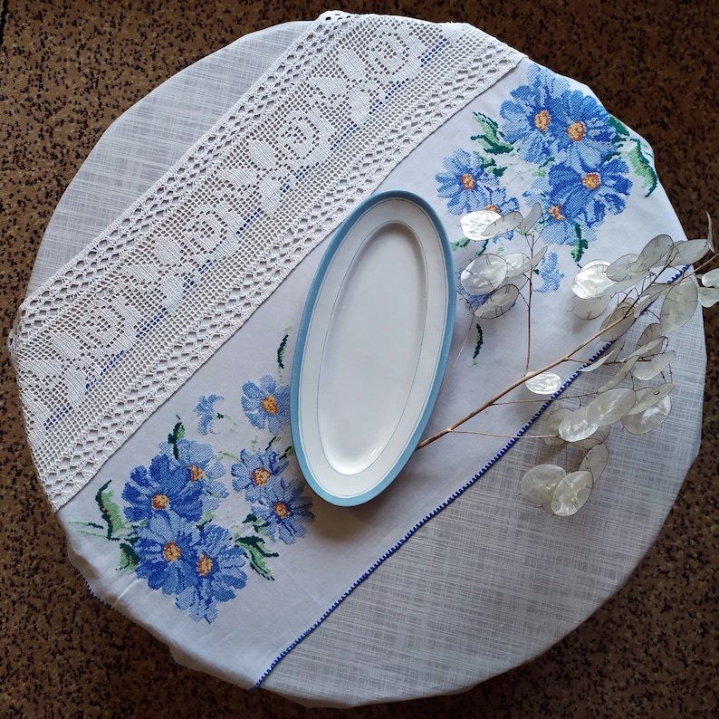 May include: A white tablecloth with a crocheted lace border and blue embroidered flowers. A white oval serving dish is placed on the tablecloth. A branch with dried seed pods is arranged on the tablecloth.