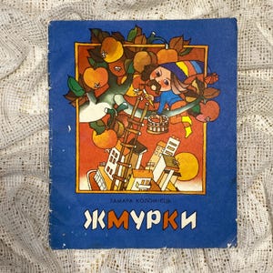 Puede incluir: Una portada de libro infantil vintage con fondo azul y una ilustración colorida de una persona en una escalera recogiendo manzanas de un árbol. El título del libro es "ЖМУРКИ" en escritura cirílica.
