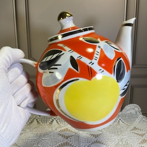 Könnte beinhalten: Eine weiße Porzellan-Teekanne mit einem gelben, orangefarbenen und schwarzen Blumendesign. Die Teekanne hat einen goldenen Rand und einen Deckel mit einem goldenen Knopf.