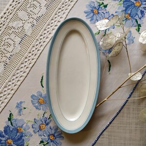 May include: A white oval serving platter with a light blue trim. The platter is on a white tablecloth with a blue floral pattern.