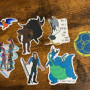 May include: A selection of vibrant stickers showcasing cartoon characters and quotes. Designs include a blue and orange bird, a horned figure, a bird with text, a couple, a dragon, and a circular design with text. The stickers are on a wooden surface.