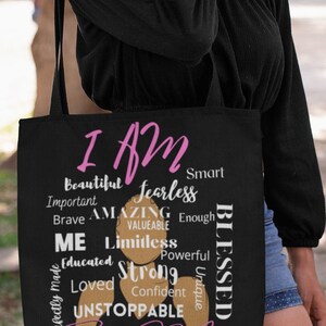 May include: Black tote bag with a pink and brown design. The bag features the text "I AM" in pink and a list of positive affirmations in black and brown. The text "Black Girl Magic" is written in pink at the bottom of the bag.