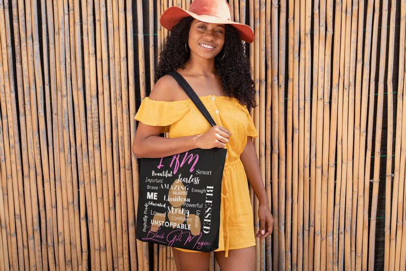 May include: A black tote bag featuring a pink and white design that reads "I am" followed by a list of positive affirmations. The bag is being held by a person wearing a yellow dress and a brown hat.