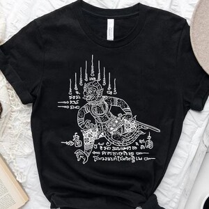 May include: Black t-shirt with a white graphic print of a Thai mythical creature. The creature is depicted in a sitting position with its hands raised. The graphic also includes Thai text.