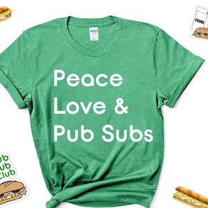 Puede incluir: Una camiseta verde con el texto "Peace Love & Pub Subs" impreso en blanco. La camiseta está doblada y se muestra sobre un fondo blanco. Hay varias imágenes de sándwiches en el fondo.