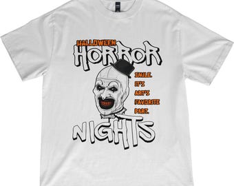 May include: White t-shirt with a Halloween theme. The shirt features a graphic of a scary clown with the words "HALLOWEEN HORROR NIGHTS" in a spooky font. The text "SMILE. IT'S ART'S FAVORITE PART" is also included.