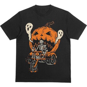 May include: Black t-shirt featuring a Halloween-themed graphic. The design includes a skeleton riding a motorcycle with a pumpkin head, flanked by two ghostly figures. The motorcycle and pumpkin are orange, with flames and smoke details.