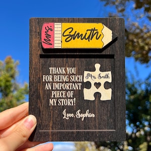 May include: A wooden teacher appreciation plaque with a pencil-shaped name tag that reads "Smith." The plaque includes a puzzle piece with "Mrs. Smith" and a heart, along with the message "Thank you for being such an important piece of my story!" and "Love, Sophia."