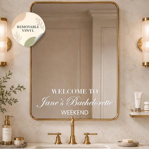 May include: A rectangular wall mirror with a thin gold frame featuring white vinyl lettering that reads WELCOME TO Jane's Bachelorette WEEKEND. A circular sticker in the top left corner displays the text REMOVABLE VINYL with botanical line art.