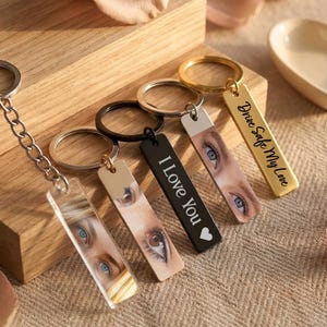 May include: A collection of keychains with rectangular pendants. Some pendants feature eye images, while others have text like "I Love You" and "Drive Safe My Love". The keychains are made of metal and acrylic, with silver, black, and gold finishes.