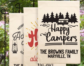 May include: Three personalized outdoor flags with a camping theme. The first flag says "Home is where you park it" with a red and yellow sunburst design. The second flag says "Happy Campers" with a black silhouette of a camper and trees. The third flag says "Adventure awaits" with a lantern design. All three flags have the text "The Browns Family, Maryville, TN, Est. 2021".