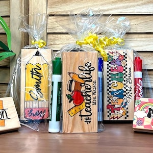 K&ouml;nnte beinhalten: Holz-Lehrerschreibtisch-Namensschilder mit farbenfrohen Designs und Lehrernamen. Eines zeigt einen Apfel und eine Kaffeetasse, ein anderes einen Bleistift und ein weiteres ein Lineal und einen Apfel. Jedes ist mit einer Klarsichtfolie und gelbem Band verpackt.