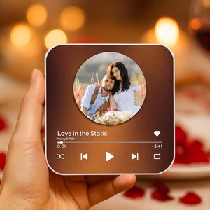 May include: A square, brown-toned device displaying a couple's photo within a circle. The device shows a music player interface with the text "Love in the Static" and playback controls. The device is held in a hand, with a blurred background of a romantic setting.