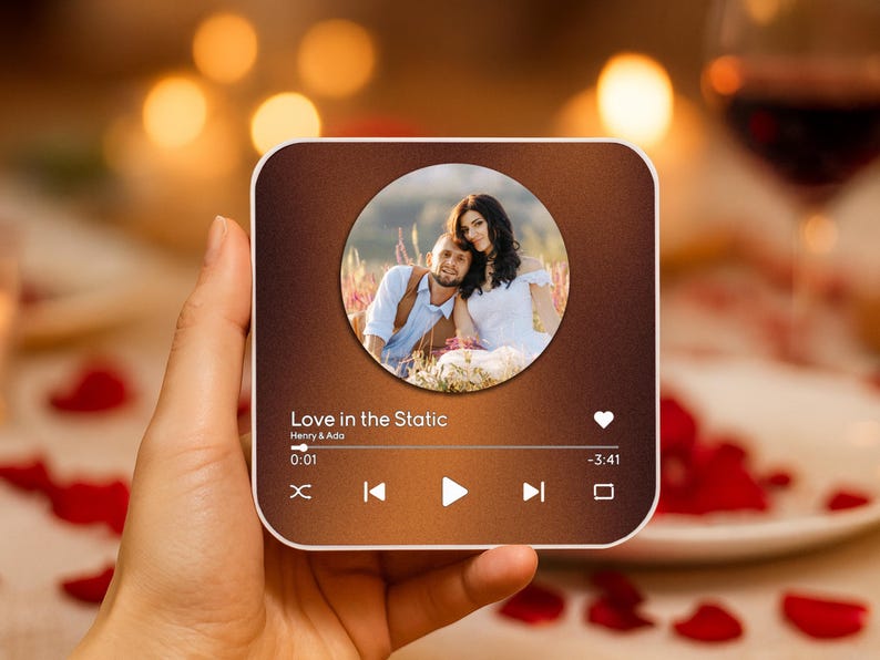 May include: A square, brown-toned device displaying a couple's photo within a circle. The device shows a music player interface with the text "Love in the Static" and playback controls. The device is held in a hand, with a blurred background of a romantic setting.