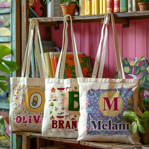 K&ouml;nnte beinhalten: Drei wei&szlig;e Canvas-Tragetaschen mit bunten Designs und personalisierten Namen. Die Taschen haben ein Blumenmuster mit dem Namen "Olivia", ein Cartoon-Muster mit dem Namen "Brand" und ein Meerjungfrauenmuster mit dem Namen "Melani".