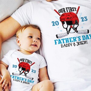 May include: A white t-shirt with a red hockey helmet graphic and the text "Our First Father's Day Daddy & Joseph 20 23". A white onesie with the same graphic and text is worn by a baby.