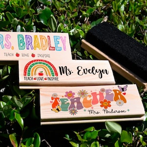 K&ouml;nnte beinhalten: Holz-Schreibtisch-Namensschilder mit farbenfrohem Text und Designs. Eines lautet "MRS. BRADLEY" mit "TEACH LOVE INSPIRE" darunter. Ein anderes sagt "Ms. Evelyn" mit einem Regenbogen. Ein drittes sagt "TEACHER" mit Blumen und "Mrs. Anderson". Ein schwarzer Radiergummi ist ebenfalls vorhanden.