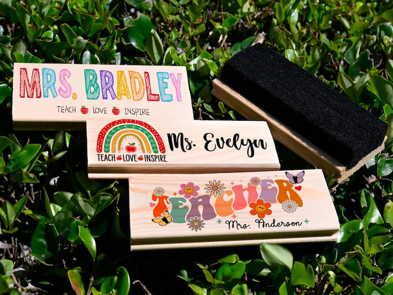 K&ouml;nnte beinhalten: Holz-Schreibtisch-Namensschilder mit farbenfrohem Text und Designs. Eines lautet "MRS. BRADLEY" mit "TEACH LOVE INSPIRE" darunter. Ein anderes sagt "Ms. Evelyn" mit einem Regenbogen. Ein drittes sagt "TEACHER" mit Blumen und "Mrs. Anderson". Ein schwarzer Radiergummi ist ebenfalls vorhanden.